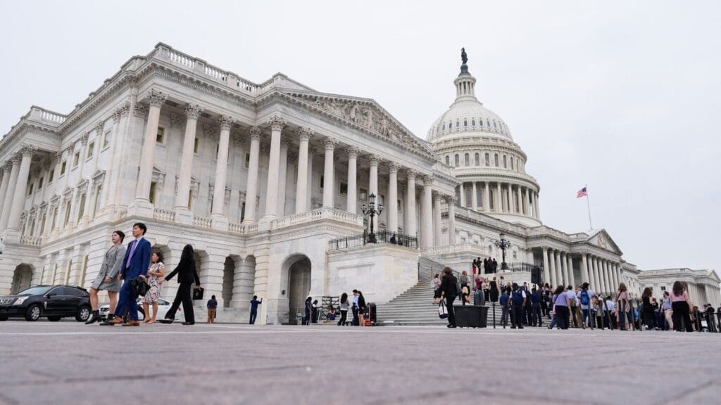 People stand in line outside the US Capitol building on a cloudy day. The Capitol’s dome and columns are visible, with the American flag flying at the top.